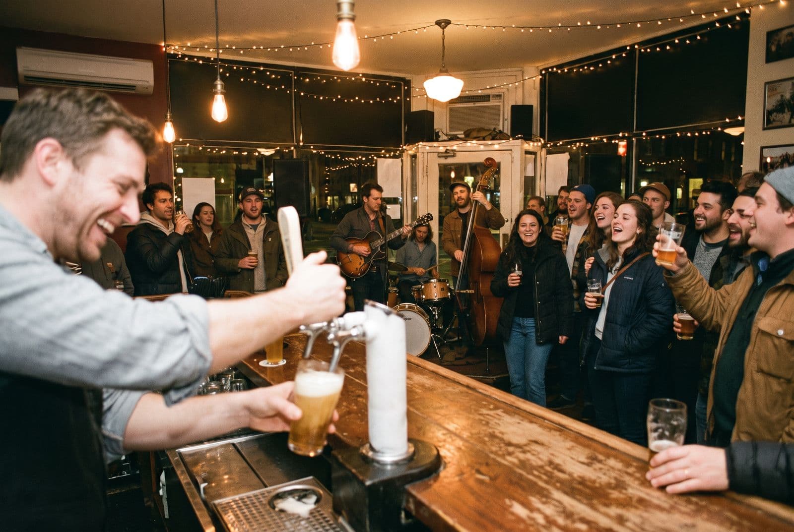 A bartender laughs while pouring a pint as a three-piece band plays to a packed, singing bar.